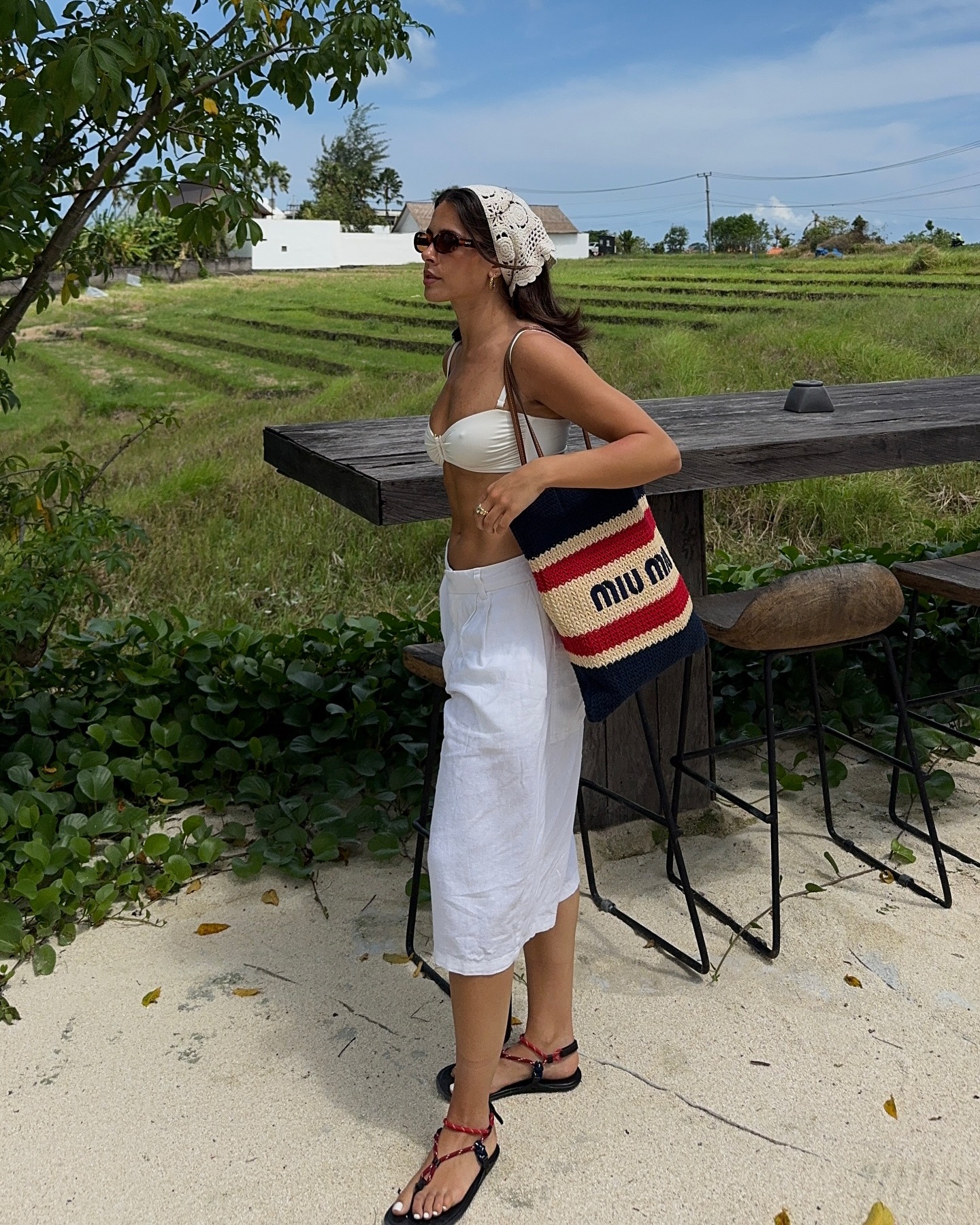 Influencer Tia Dewitt wears a MiuMiu knit bag, linen trousers, a crochet hair scarf, and rope sandals.