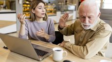 An adult daughter and her father have a disagreement while in a coffee shop looking at a laptop.