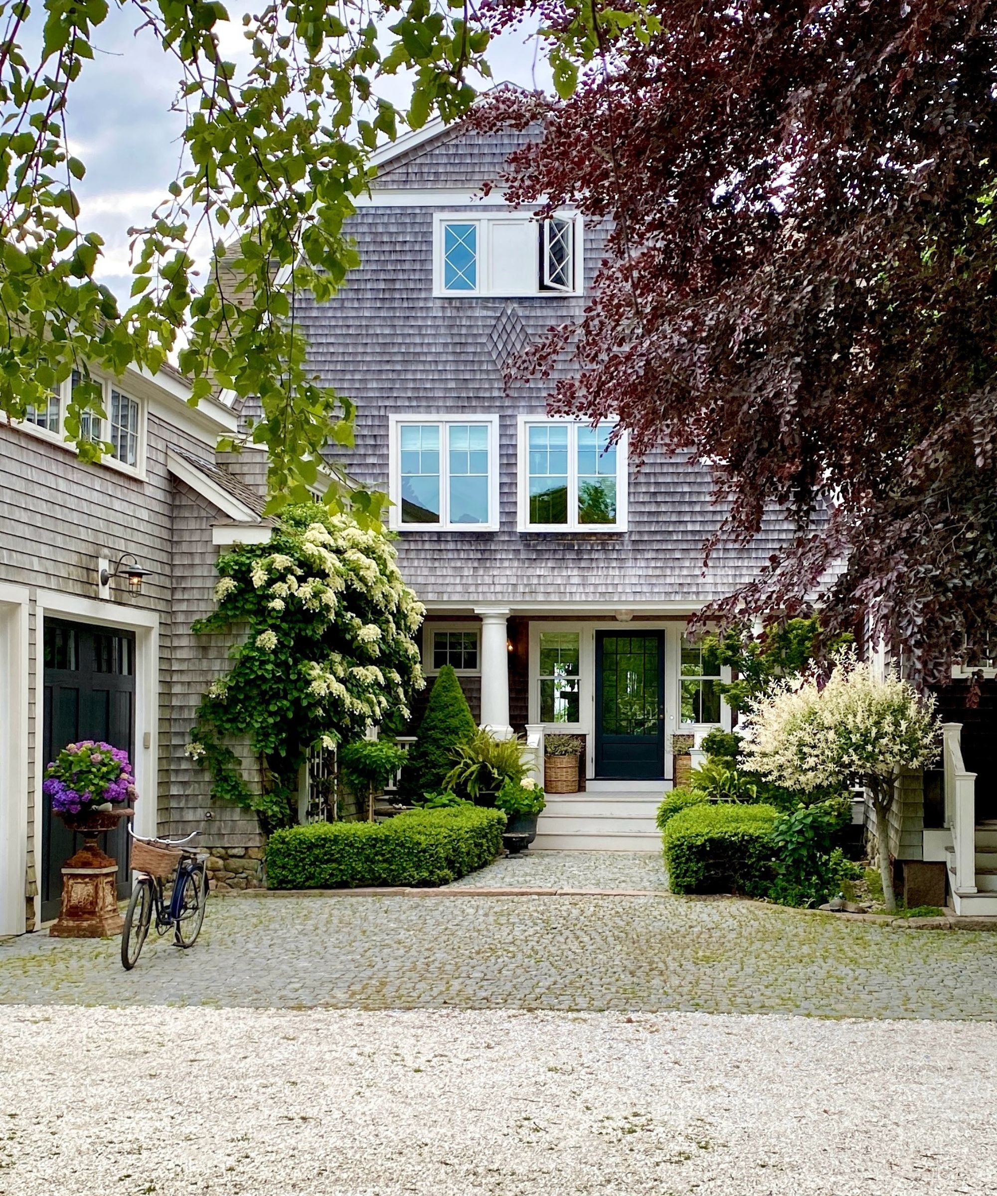Shingle-style house with a pillared entrance, a cobbled pathway, hedges, a garage with a bicycle outside, hydrangeas, and potted plants
