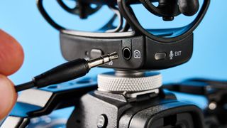 a black shotgun microphone by joby called the wavo pro is photographed by itself against a blue background, showing its muff, dead cat, attachment foot, and connection ports