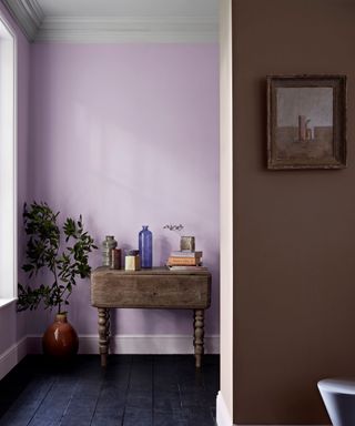 Corner of room, mauve painted walls, small wooden console table decorated with ornaments, black wooden flooring, other brown painted wall