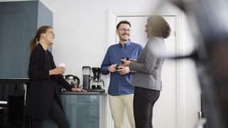 Three colleagues stood talking next to a coffee machine in an office, to show working in the office compared to working from home. The shot is low, taken from seat height with an object partially obscuring the frame on the right-hand side.