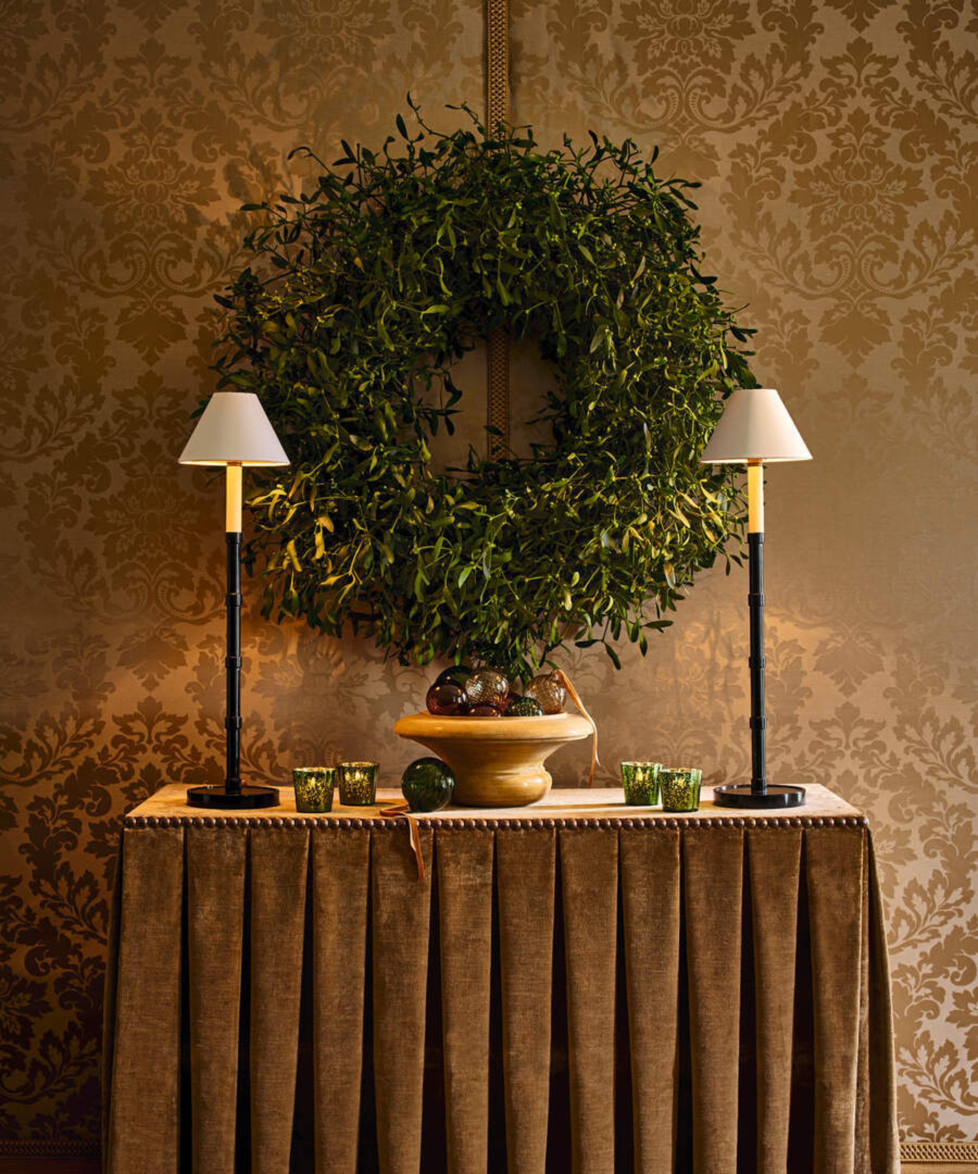 a large green wreath hanging on a wall behind a table with two lamps and a bowl of ornaments