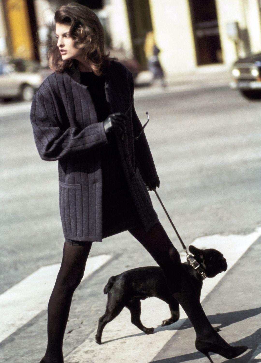 Model Linda Evangelista, crossing a street with a dog, wearing a long gray wool flannel jacket with vertical black stripes woven in, over a short black wool skirt and top, with matching stockings and leather gloves by Azzedine Alaia. Shoes from Alaia by Diego Della Valle.