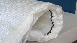 A mattress wrapped in plastic slowly unrolling and expanding on bed base