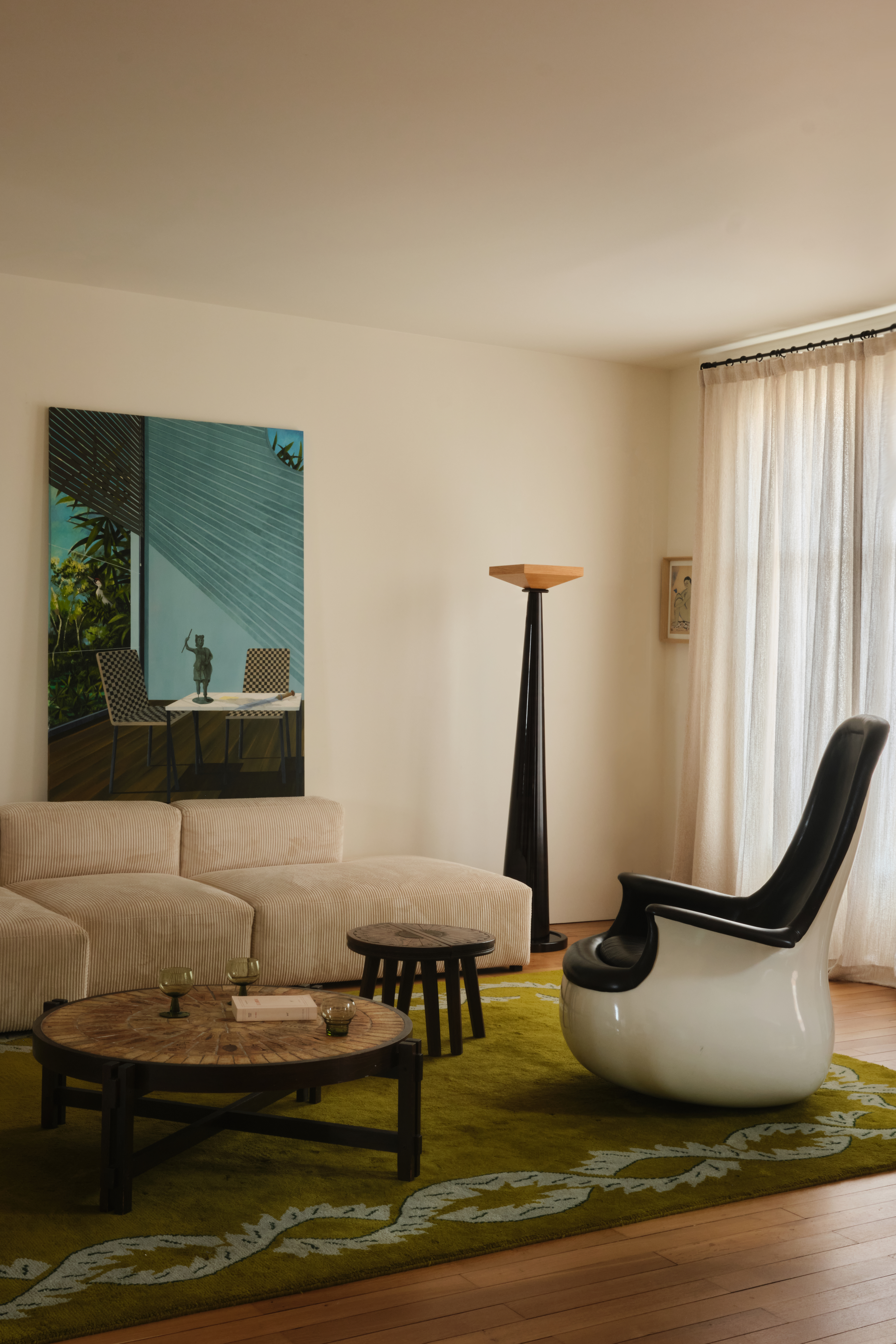 A modular sofa in beige, a sculptural armchair in white and black lacquered resin, a standing lamp, and a figurative artwork with a blue background sit in a neutrally painted living room.