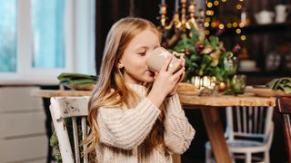 A young girl drinks from a mug while sitting by a Christmas tree