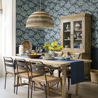 Dining room with blue patterned wallpaper on all walls with wooden furniture and woven light shade