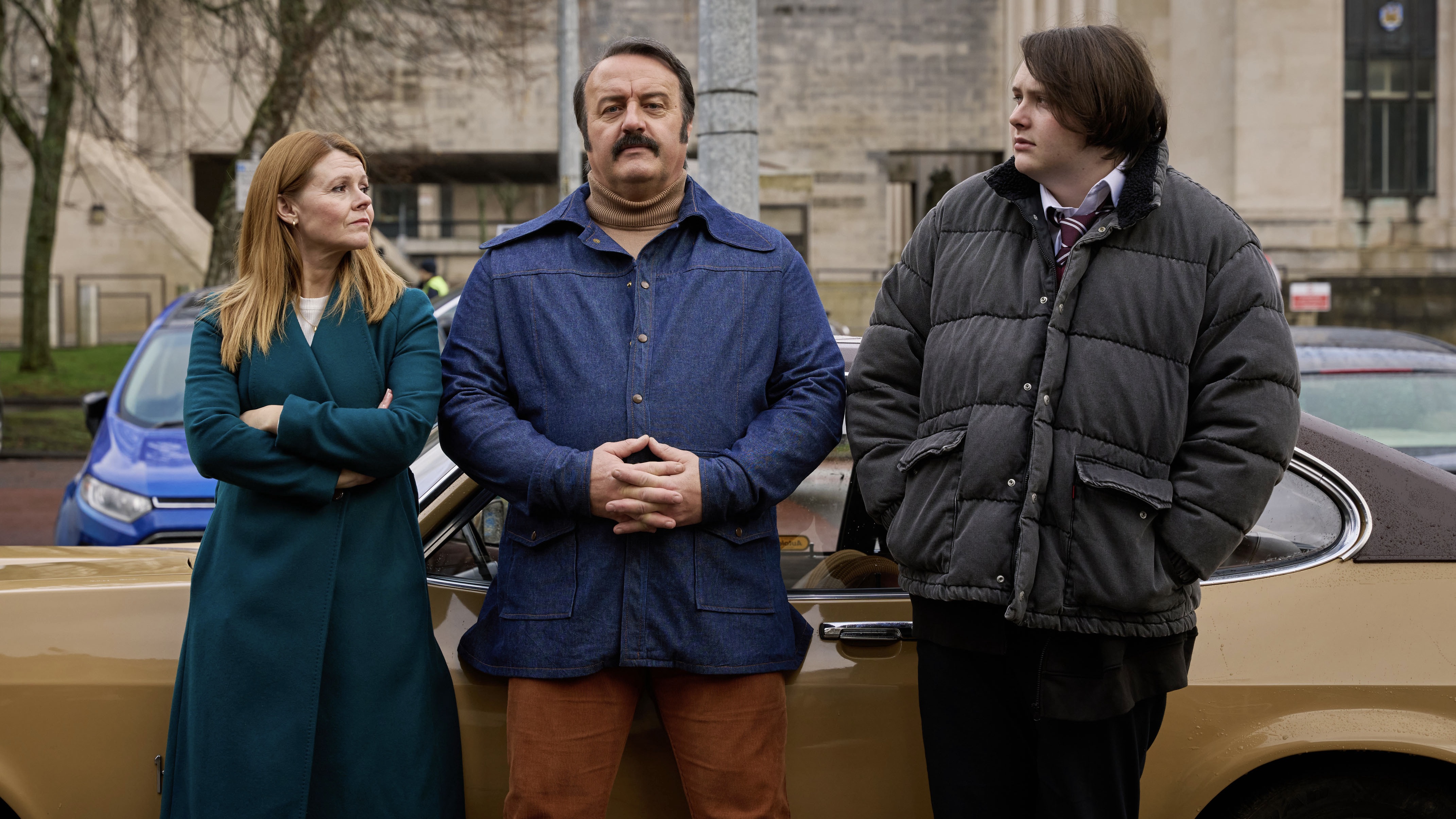 Sian Gibson in a green coat as Mel stands next to Mike Bubbins in a denim jacket as Tony Mammoth and Joel Davison in a black coat as Theo alongside a beige Ford in Mammoth.