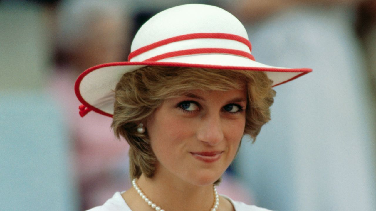 A headshot of Princess Diana smirking wearing a white hat with red trim and pearls