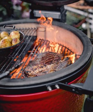 A steak cooking flame-grilled style on a BBQ with a side of potatoes