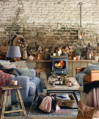 Living room with a lit woodburner, natural stone wall with logs piled on a wooden shelf, armchairs covered with cushions and throws.