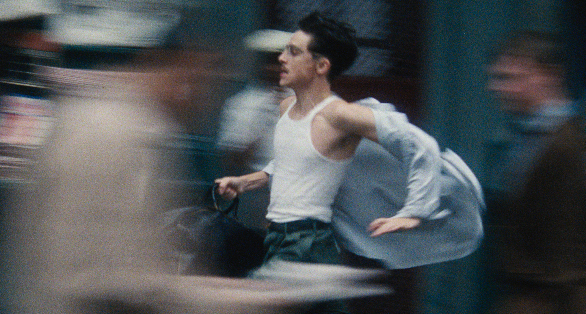 A man (Timothee Chalamet as Marty Mauser) wears a white tank top and carries a black bag, with a loose button-up shirt blowing behind him as he runs. The crowd around him is blurred, in a still from 'Marty Supreme.'