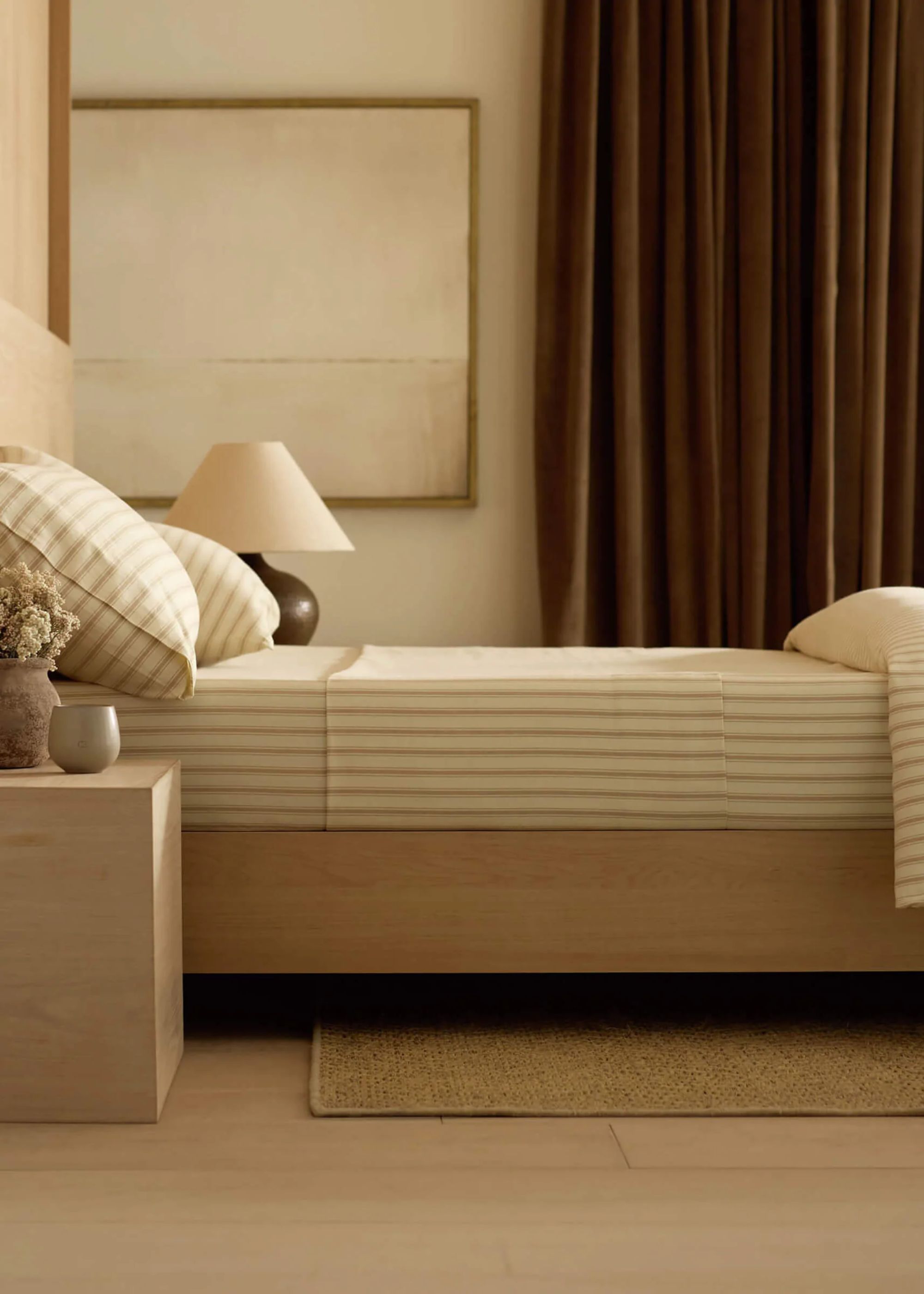 A bedroom with striped bedding, a wood side table, a table lamp, and a minimalist painting