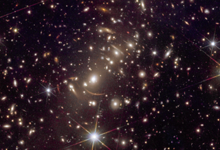 A stunning view of the dark background of space with a slightly purplish-brown glint. Tons of larger yellowish speckles are seen and many smears indicated gravitationally lensed galaxies are visible in the image as well, forming faint slices of rings in the center.