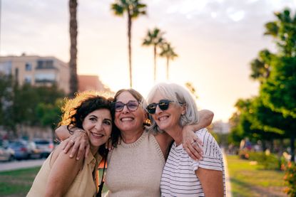 Three lifelong friends are embracing and smiling happily on the street at sunset, in backlight. The sunlight behind them outlines their silhouettes, highlighting their joy and the deep bond of friendship they share.