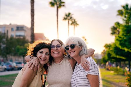Three lifelong friends are embracing and smiling happily on the street at sunset, in backlight. The sunlight behind them outlines their silhouettes, highlighting their joy and the deep bond of friendship they share.