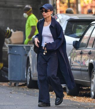 Zoe Kravitz in NYC wearing a navy baseball cap, sunglasses, a white tank top, blue long coat, navy jeans, and the Row Zipped boots in black.