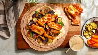 Halloumi and roasted red onion, courgette and bell peppers arranged on a wrap, next to a plate of roasted vegetables and a bowl of hummus