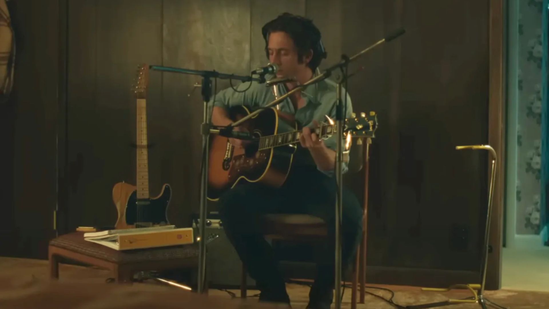 an image of jeremy allen white playing bruce springsteen in the movie Deliver me from Nowhere. He is sitting on a chair in a recording studio. He is singing into a mic with headphones on and playing guitar