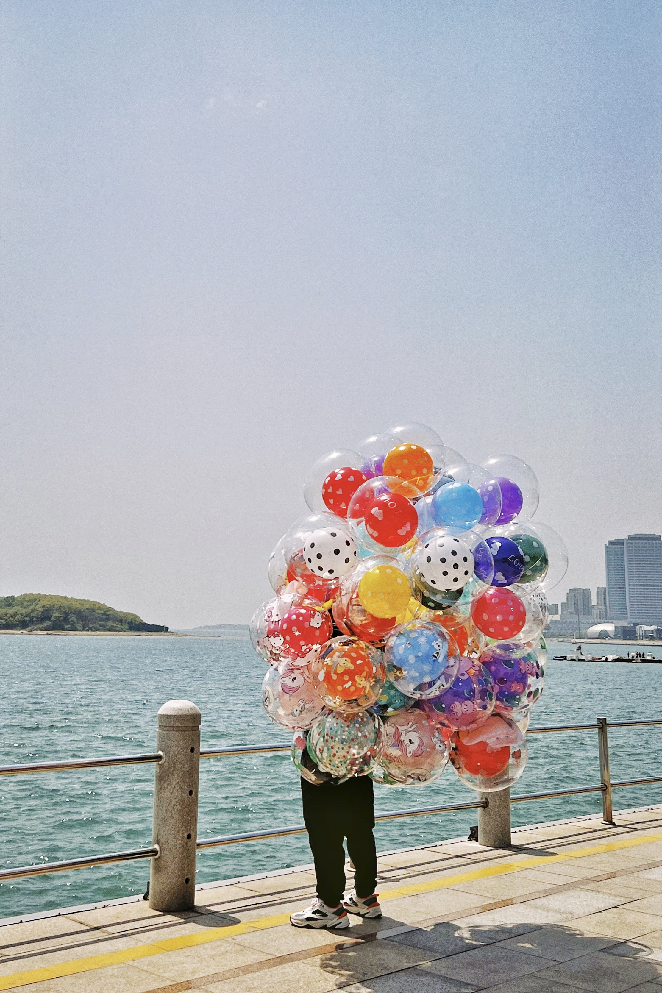 A man with several colorful balloons stands in front of a city river