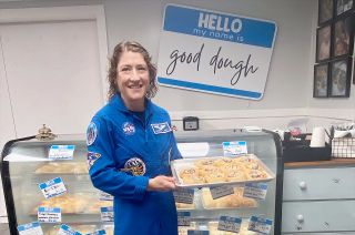 an astronaut in a blue flight suit holds a tray of pastries in a bakery
