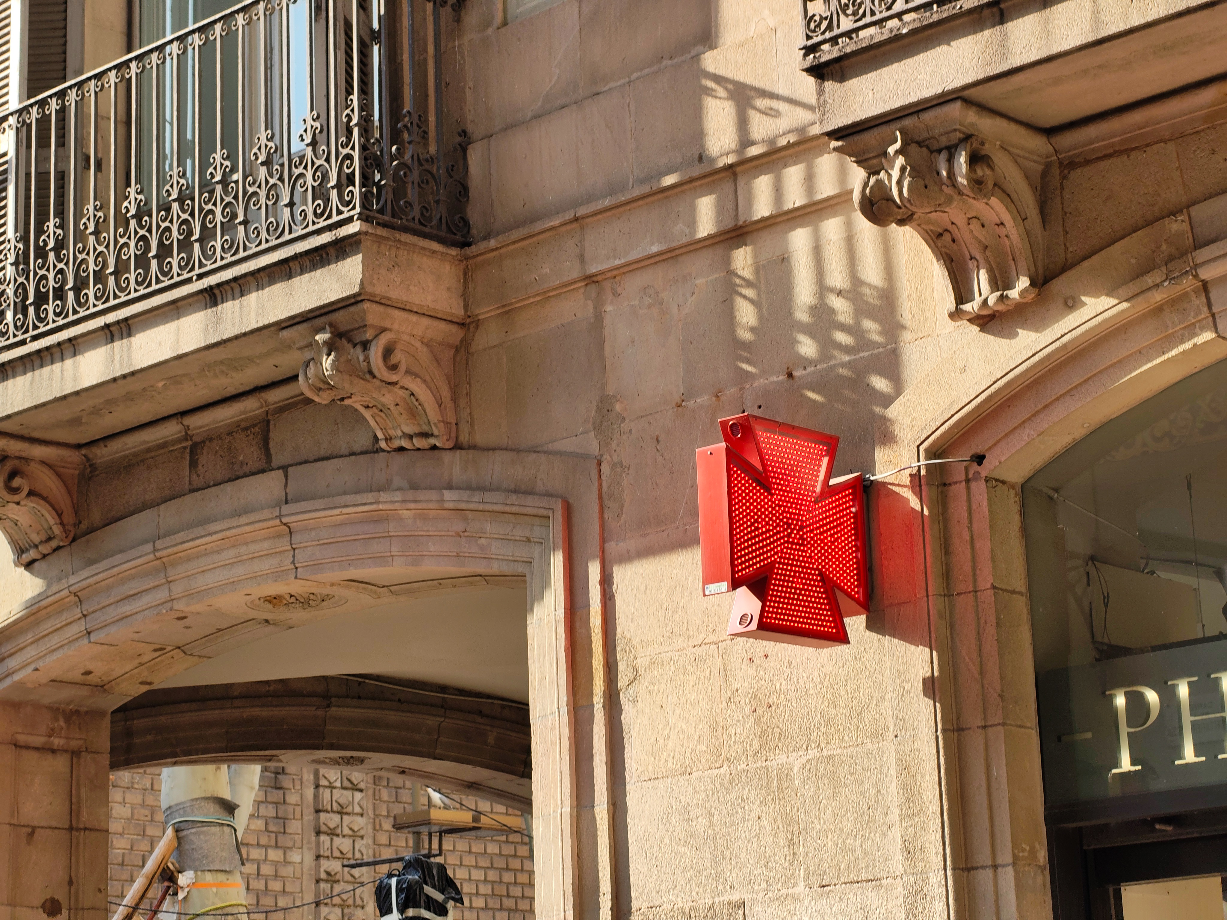 Red illuminated pharmacy cross sign mounted on a stone building facade in warm afternoon light, photographed with the Nothing Phone (4a).