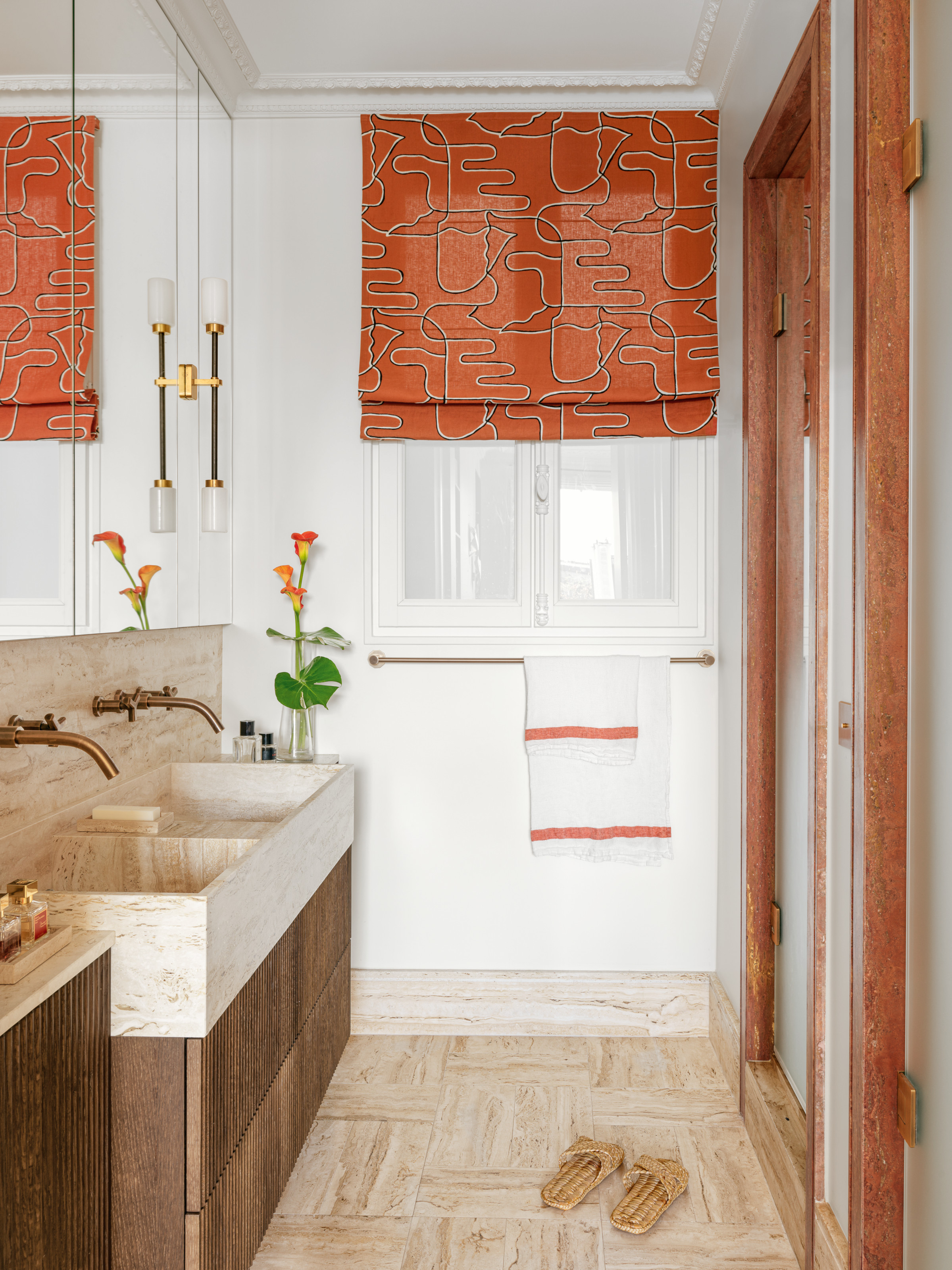 Bathroom with wood and beige stone vanity, rust marble surround to shower, and orange patterned Roman blind