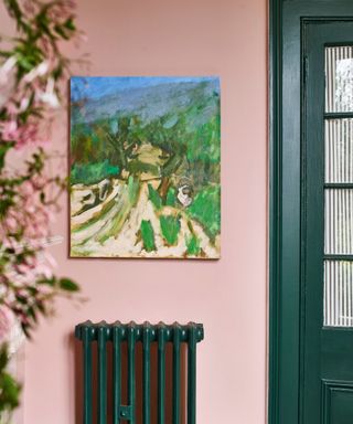 A pink entryway with a green door and radiator, a painting of a countryside scene, and pink flowers to the left