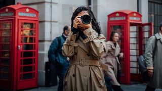 A woman in a beige trench coat stands among pedestrians near iconic red phone booths in London
