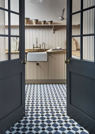 A cream kitchen with geometric blue and white tiled floor, black painted doors, a butler sink and open wooden shelving.