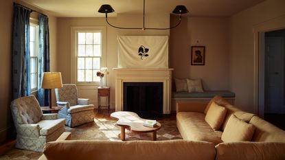 Cozy living room with a cream fireplace, a unique organic-shaped wood coffee table, two patterned armchairs, and a large white banner with a black leaf design over the mantel