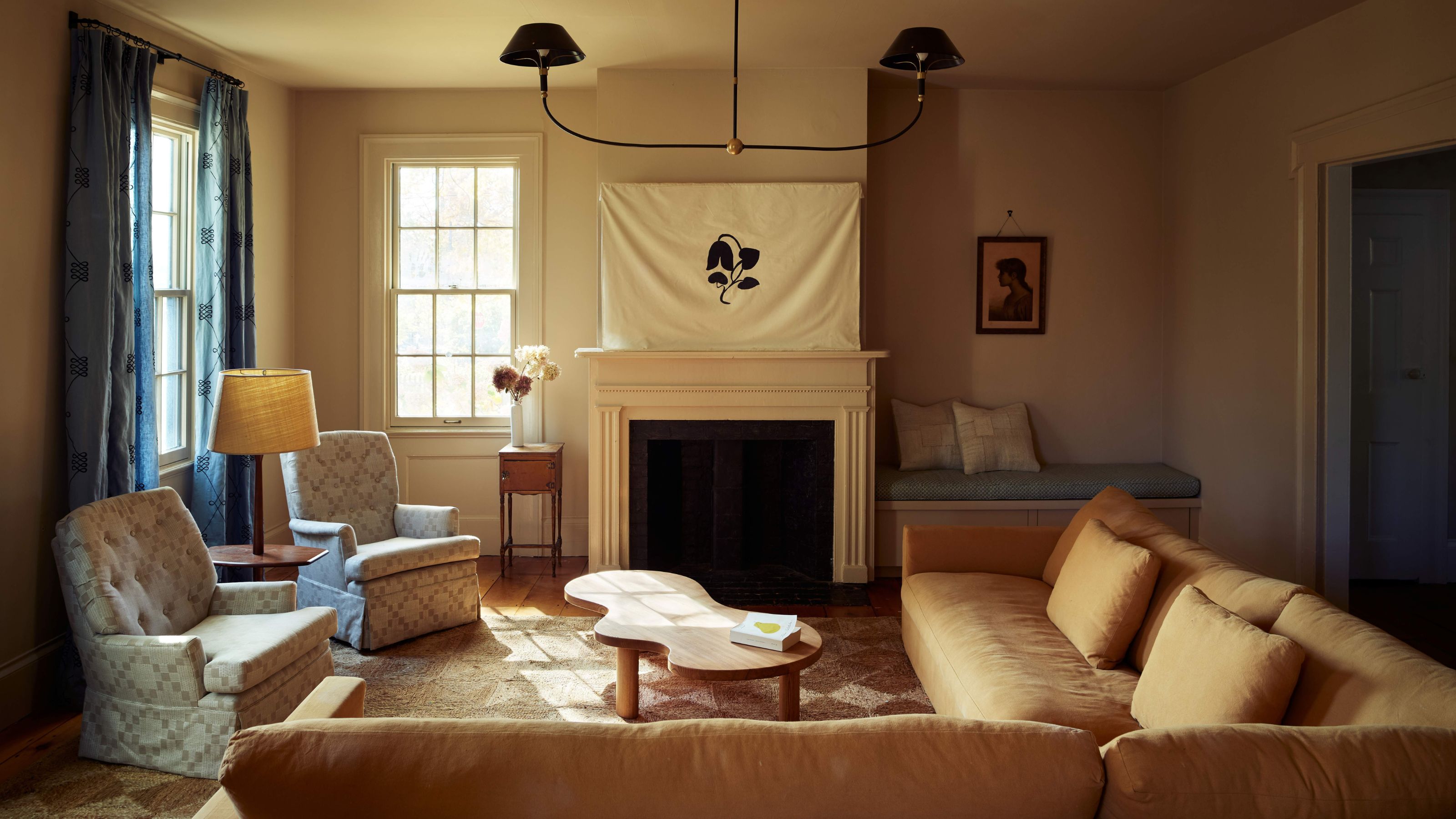 Cozy living room with a cream fireplace, a unique organic-shaped wood coffee table, two patterned armchairs, and a large white banner with a black leaf design over the mantel