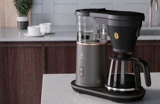 En svart Electrolux Explore 7 står på en grå köksö, med en köksbänk och två vita kaffekoppar i bakgrunden.