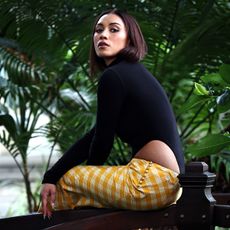 Luciane Buchanan photo taken during a press day for her new show Chief of War. She is wearing a custom look by Hawaiian designer Rocket Ahuna consisting of a black high neck, long-sleeve top that cuts up on the side and a yellow and white gingham maxi skirt. 