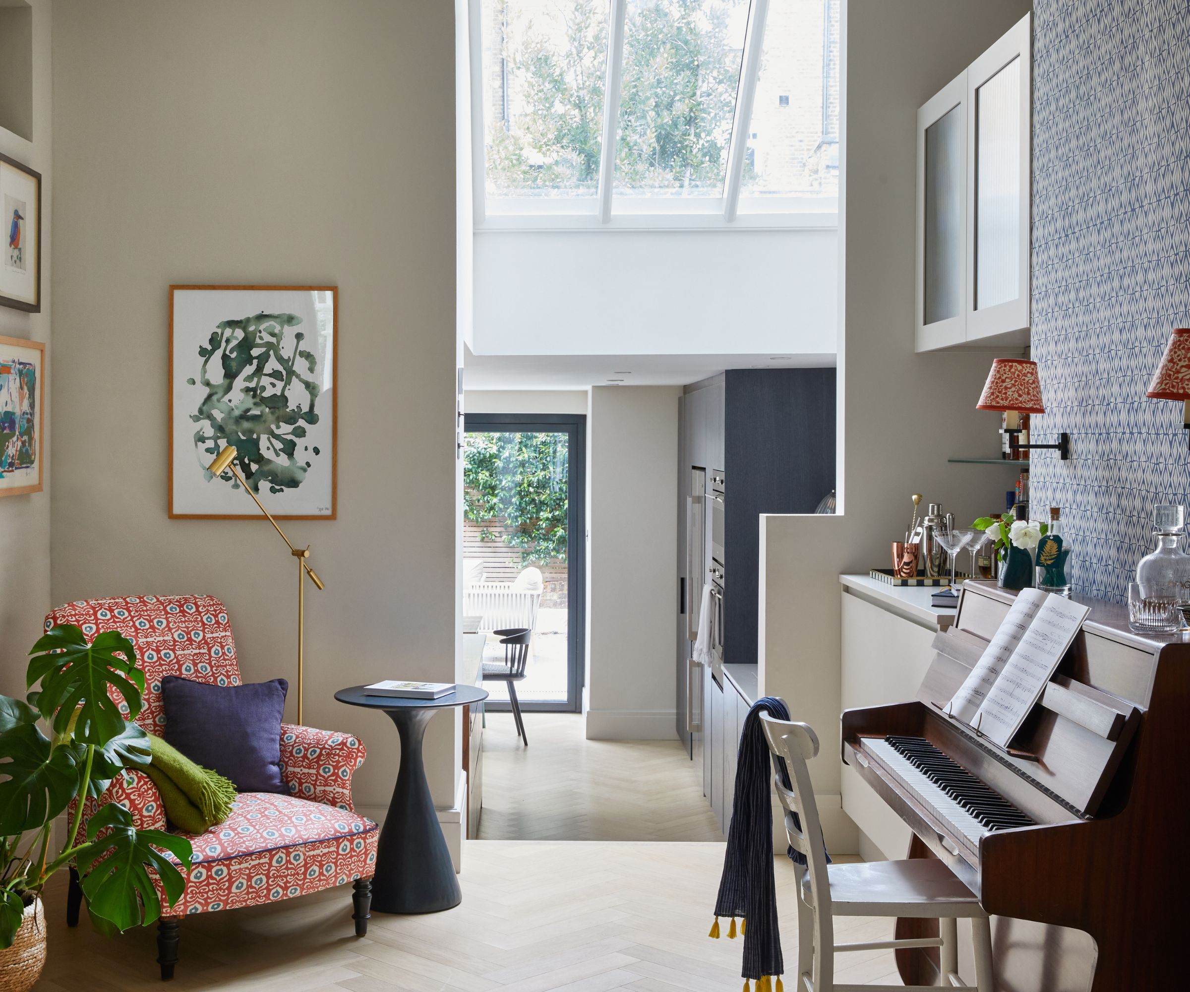 Open plan living room with piano, two red sconces on the wall above it, a reading corner with a red armchair and a gold floor lamp with artwork above, leading into an open plan kitchen with a skylight