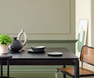 A two toned green dining room with a darker shade on the bottom of the wall and al ighter shade on top, a wooden dining table and one chair.