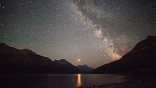 A glowing strip of the Milky Way galaxy is seen in a dark gray and purple starry night sky overlooking a lake surrounded by silhouetted jagged peaks on all sides with a glowing bright dot in the center of the image casting a yellow column of light on the dark water