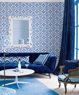 A blue living room scheme with a dark blue sofa and drapes, round white coffee table and mirror, and wallpaper in the style of Portuguese blue and white tiles