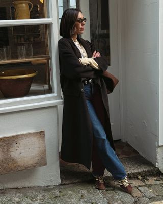 British style influencer Jessica Skye wearing a winter outfit with oval sunglasses, a long brown coat, cream button down cardigan, Khaite studded belt, brown suede bag, dark-wash jeans, and brown suede Adidas sneakers