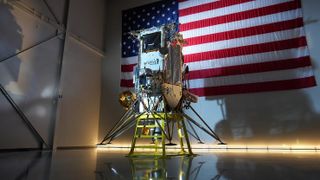 a tall, silver moon lander sits on a stage in front of a large american flag