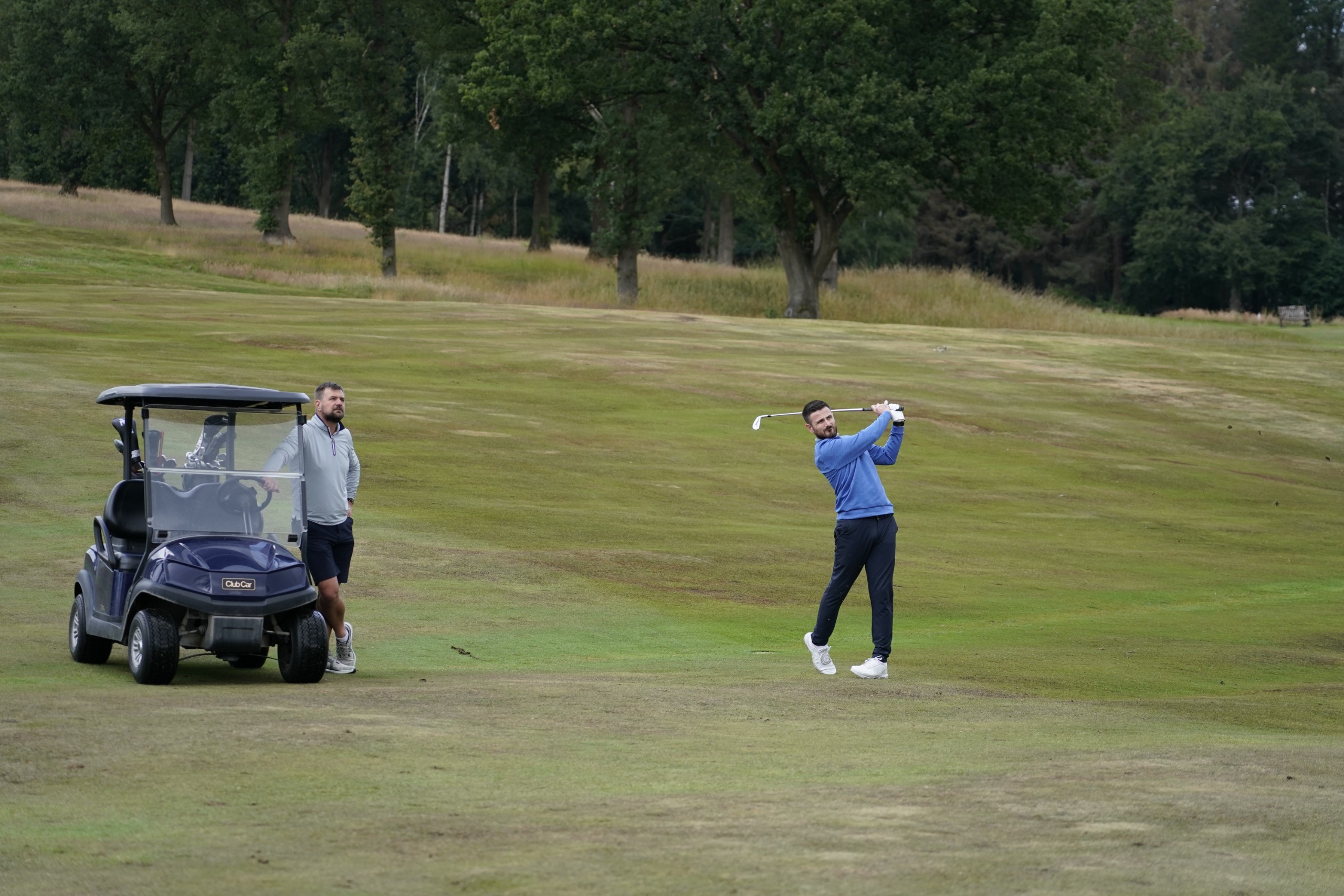 Baz Plummer hitting an iron shot from the fairway while being watched by his coach, Alex James