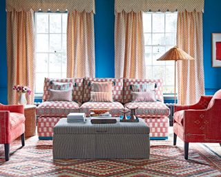 Blue living room with yellow curtains, pink sofa and matching armchairs, patterned rug, striped ottoman.