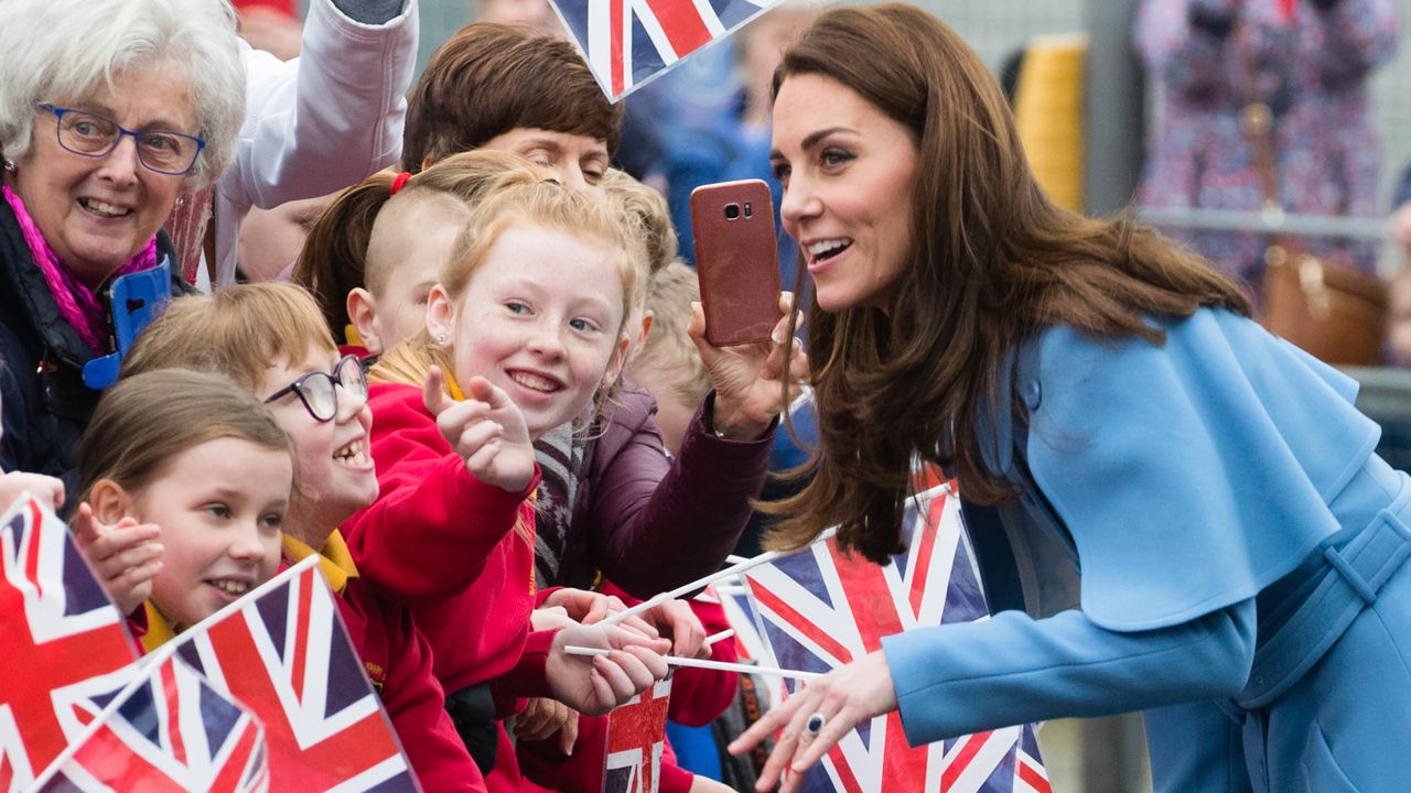 Kate Middleton wearing a blue cape coat leaning forward to talk to kids waving Union Jack flags