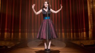 Rachel Brosnahan standing on stage behind a microphone holding her arms up and addressing the audience.