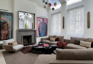 modern living room with fireplace with large artwork hung on either side and a mirror hanging over the fire, two windows with sheer curtains, a large L-shaped brown sofa with a nesting coffee table and modern white and timber armchair