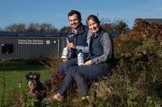 A man and a woman sit on a grassy bank with a dog holding bottles of gin