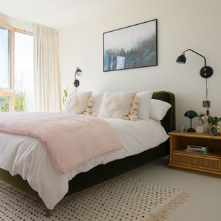 A master bedroom with cream curtains, green velvet bed frame, white bedding, cream cushions with tassels, black framed wall art and pink throw
