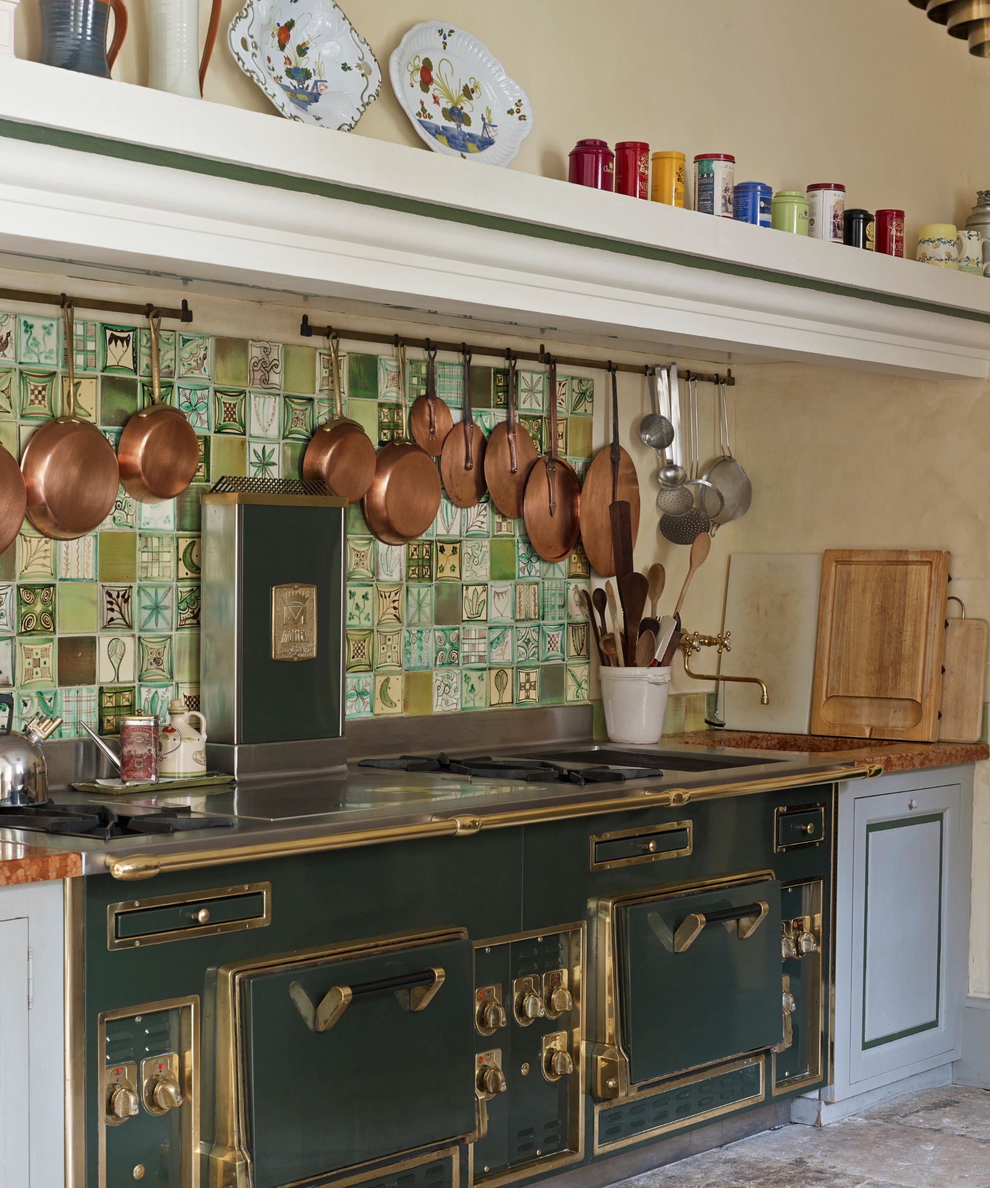 Kitchen with green tiles, green vintage stove, copper pans hanging up, ceramics on shelf