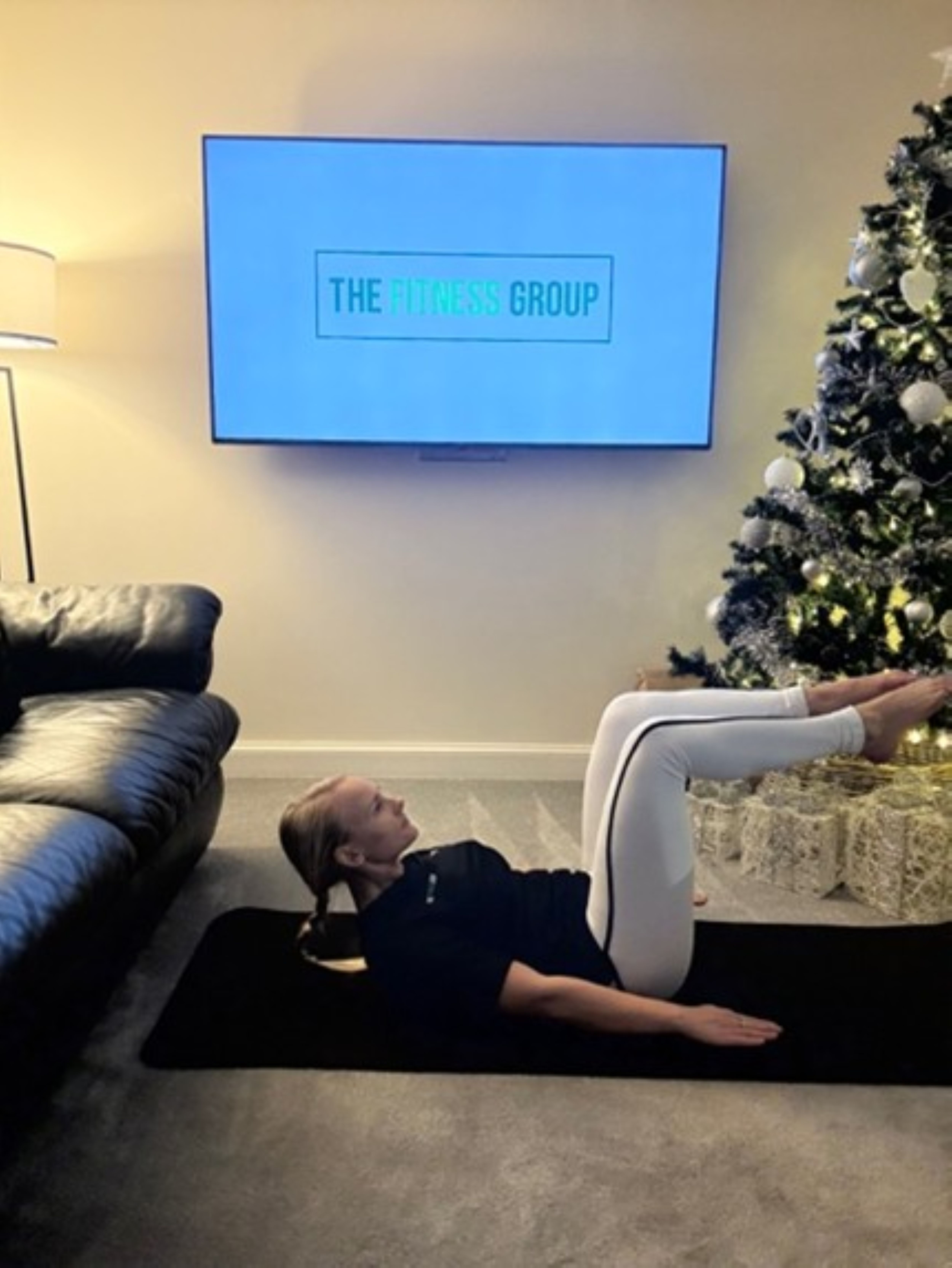 Woman demonstrates a Pilates exercise between a sofa and a Christmas tree. A mounted TV in the background reads "The Fitness Group"